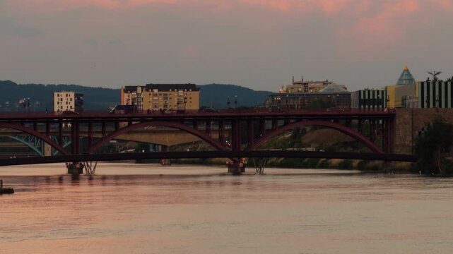 The Old Bridge spans the Drava River in Maribor as warm sunset light reflects on the water, with modern city buildings rising behind the steel arches.