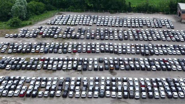 Automotive import and export distribution yard stores thousands of passenger vehicles in organized inventory rows within a large logistics terminal supporting global supply chains, drone pullback.