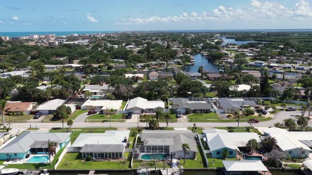 Waterfront homes line canal with private boat docks plus swimming pools, palm-lined street stretching toward distant high-rises near ocean in Cocoa Beach, Florida area. Aerial drone flyover shot