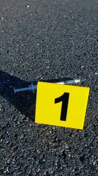 Bright yellow evidence marker with the number one next to a used syringe on the asphalt, marking a piece of evidence at an outdoor crime scene related to drug use or overdose