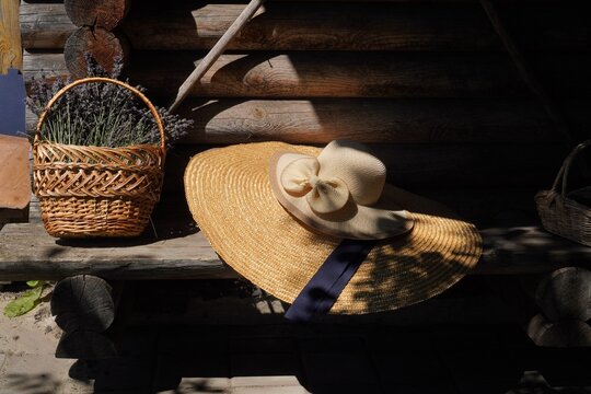 Straw hat resting on a wooden bench beside a basket filled with fresh lavender. The setting includes a rustic wooden backdrop, evoking a serene and tranquil atmosphere