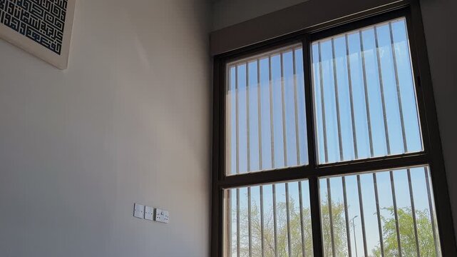 A dark roller blind is being pulled down to cover a barred window in a modern home interior. Bright daylight shines through the glass, illuminating the white wall and geometric artwork.