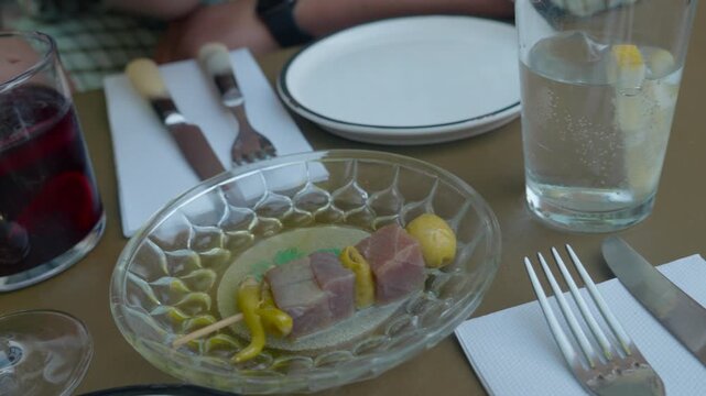 Close up of a woman's hand taking a tapas skewer of marinated tuna, olives and pickled peppers from a glass plate on a set table. Authentic Spanish appetizer moment.