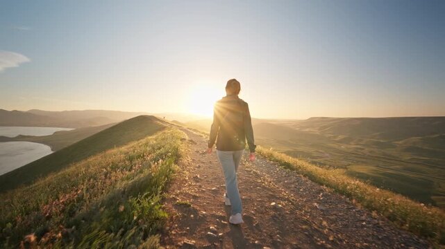 Young woman hiking along a dirt path on a hill overlooking a beautiful sea bay at sunset. Cinematic slow motion steadicam follow shot footage for active lifestyle, summer travel and freedom video.