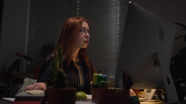 Young adult Caucasian woman with glasses working late on computer in dark office. Footage is suitable for corporate training, remote work promotions, or productivity campaigns