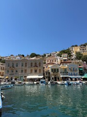 Obraz premium Colorful Symi harbor houses by the sea, turquoise Aegean water, traditional Greek island architecture, bright summer sunlight, peaceful coastal view