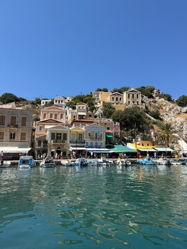 Colorful Symi harbor houses by the sea, turquoise Aegean water, traditional Greek island architecture, bright summer sunlight, peaceful coastal view