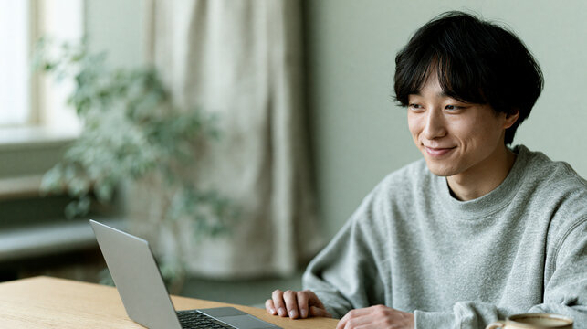 明るい室内でノートパソコンに向かい微笑む二十代の男性 ｜ ナチュラルな背景