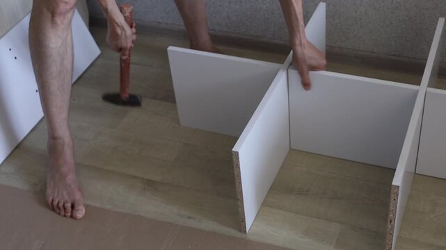 Man assembles furniture using hammer and wooden panels on floor indoors