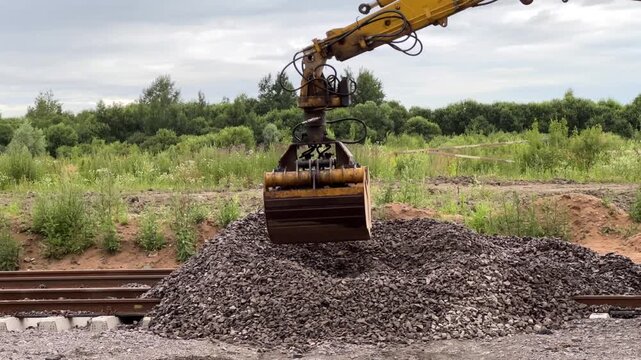 An excavator with a bucket pours gravel between railway tracks during construction. Railway construction, heavy machinery, track maintenance.