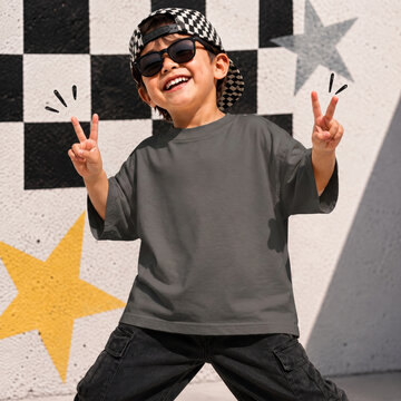 Boy in graphite tee, with sunglasses posing by colorful wall. T-shirt front side view. Kids short sleeve t-shirt template. Children shirt mockup summer youth outdoors