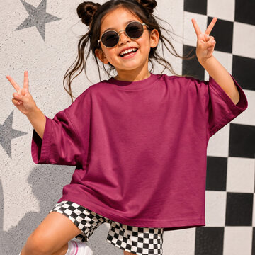 Girl in berry tee, with sunglasses posing by colorful wall. T-shirt front side view. Kids short sleeve t-shirt template. Children shirt mockup summer youth outdoors