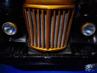 Front view of a restored retro vehicle featuring a distinctive gold vertical bar grille, mesh radiator screen, and classic circular headlamps. © anas