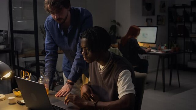 Medium full shot of Caucasian young adult man mentoring African American young adult man typing on laptop in office at night. Footage for corporate training videos or business teamwork presentations