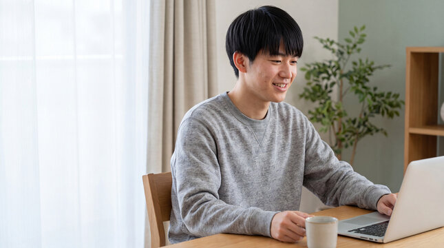 明るい室内でパソコン作業をしながら微笑む若い男性 ｜ 自然光の入る部屋