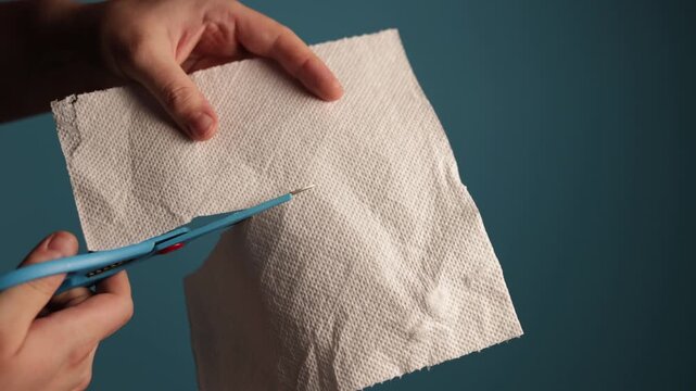 Hands cutting a paper towel with blue scissors on a teal background