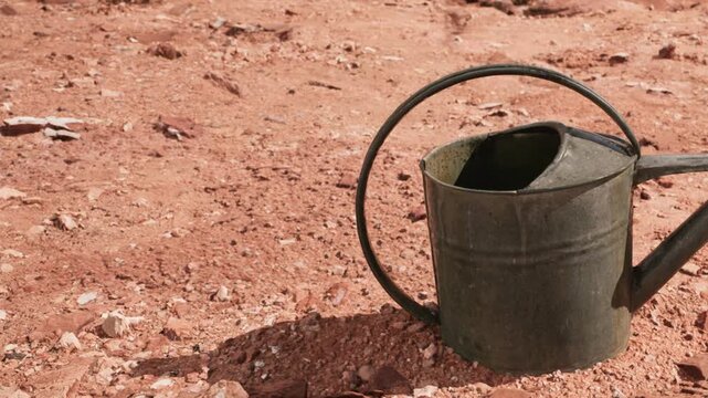 A metal watering can is placed on dry ground in a barren landscape. The soil appears cracked and lacks vegetation. The scene shows the harshness of the environment and the need for water.