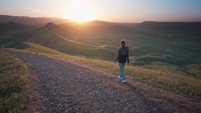 Young woman hiking along a dirt path on a hill overlooking a beautiful sea bay at sunset. Cinematic steadicam follow shot footage for active lifestyle, summer travel and freedom video.	