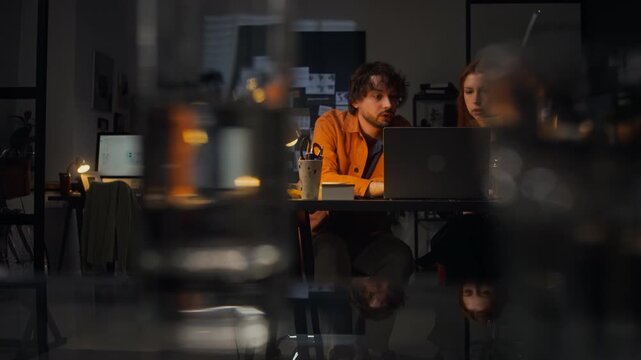 Wide shot of Caucasian young adult man and Caucasian young adult woman working late in office, looking at laptop, copy space. Clip perfect for corporate teamwork and startup business marketing videos