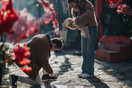 A man kneels to tie a woman's sneaker while they laugh and interact as friends. The casual outdoor scene on a sunlit cobblestone street shows a playful moment between companions.