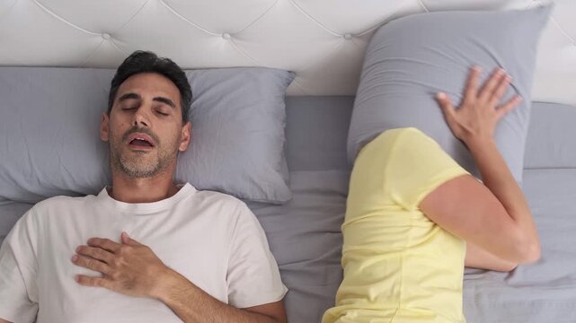 Woman covering head with pillow while partner snores in bed
