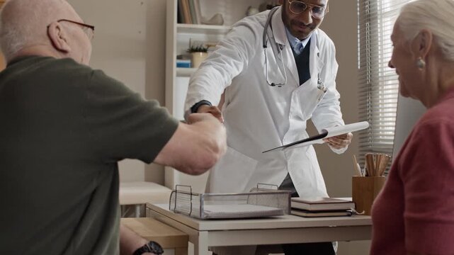 Indian male doctor shaking hands with senior Caucasian couple during medical consultation in clinic. Clip is perfect for healthcare services, medical insurance, and senior care advertising