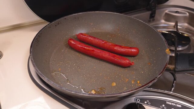 Two red sausages sizzling in hot oil inside a non-stick frying pan on a gas stove, cooking a simple meal in a recreational vehicle kitchen during a road trip or camping adventure