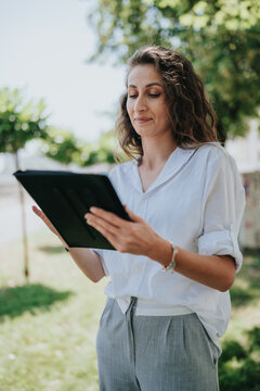 Young businesswoman standing outdoors reading a tablet with a focused expression. Casual professional scene showing remote work and digital productivity in a park setting.