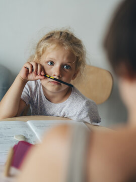 A girl with big, intelligent blue eyes looks at her mother and listens attentively while doing her homework. She holds a pencil to her face.