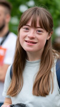 Young woman with Down syndrome smiles while holding a camera, surrounded by blurred figures and greenery in an outdoor setting during a social gathering