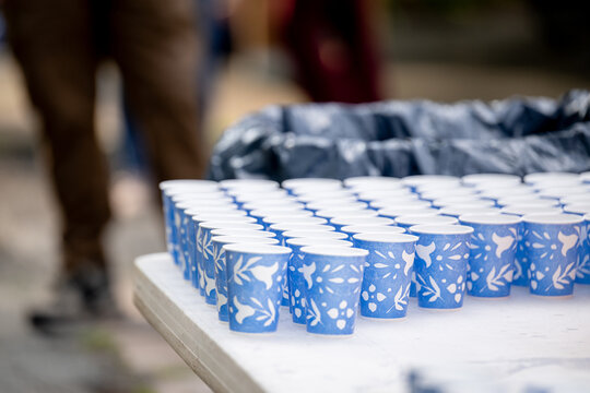 Neat rows of blue patterned paper cups wait on a white table at an event hydration station, poured and lined up before runners arrive, as passersby walk behind.