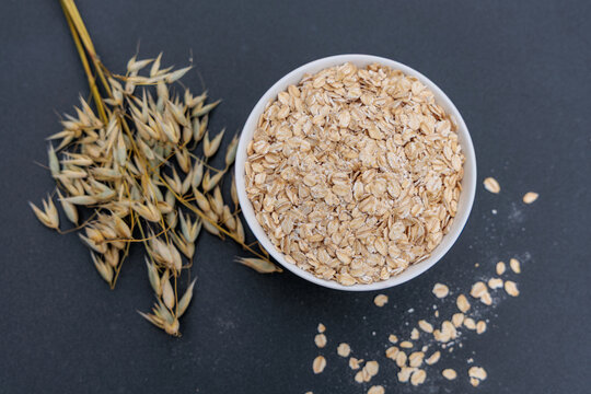 Rolled oats in white bowl with dry oat ears and spilled flakes on dark background, healthy breakfast ingredients, top view flat lay