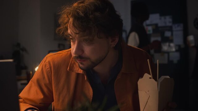 Caucasian young adult man eating takeout food from box with chopsticks while looking at laptop screen in dark office at night. Footage represents corporate overtime or food delivery advertisements
