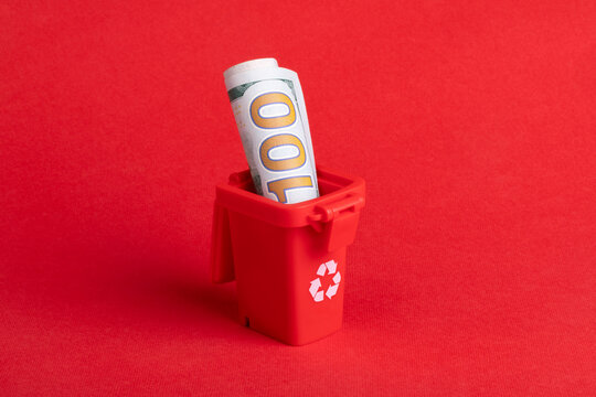 Rolled hundred dollar bill placed in a small red recycling bin on a solid red background, symbolizing financial waste or sustainability concepts.