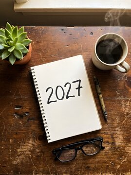 2027 written in a spiral notebook on a wooden desk with a cup of coffee and a green succulent plant next to a pair of black reading glasses for new year planning and goal setting