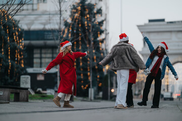 Fototapeta premium Three girls wearing Santa hats laugh and dance together on a city street decorated with lights. The festive friends celebrate a winter evening under holiday lights and archways.