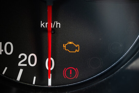 Close up view of an illuminated check engine light and hand break on warning signal on a black car dashboard speedometer scale.