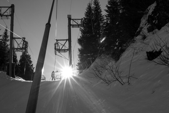 A skier's perspective from a ski lift chair captures bright sun flaring through the lift cables and pylon with silhouetted figures ahead.