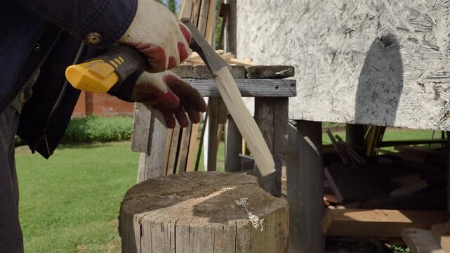 A gloved person splitting a thin wooden block with an axe on a tree stump in a sunny outdoor setting, suitable for DIY or homesteading content.