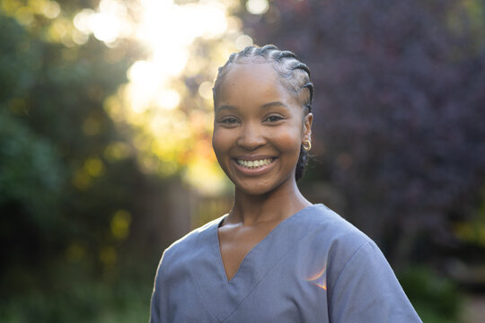 African woman standing in garden smiling wearing blue-gray scrub cornrows hoops sunlit bokeh