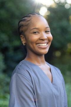 African American woman standing in park wearing gray scrubs showing cornrows and gold hoops smiling