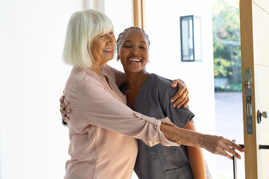 African American caregiver in scrubs and senior woman in pink hugging at front door, holding handle