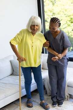 African American caregiver assisting senior woman in scrubs in living room with cane