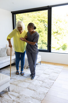 Senior woman and African American caregiver in scrubs walking on rug at home holding wooden cane