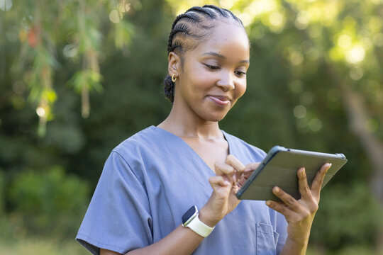 African American woman holding tablet in case tapping screen wearing light blue scrubs in park