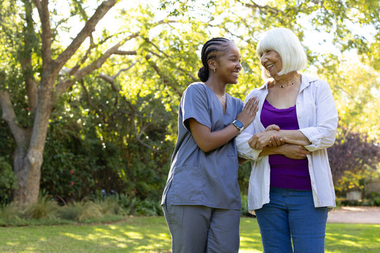 African American caregiver holding senior woman's forearm, smiling on lawn under tree in scrubs