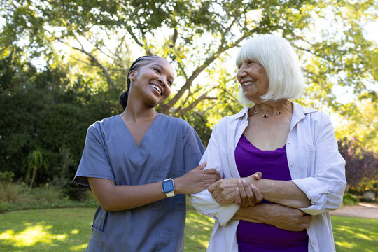 Female carer in scrubs checking watch, linking arms with female senior with necklace in garden