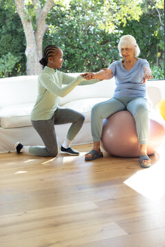 Diverse women practicing balance, caregiver holding senior's hands on pink exercise ball at home