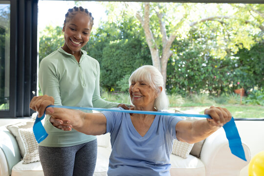Senior woman and African American woman trainer practicing resistance band exercise on sofa in tops