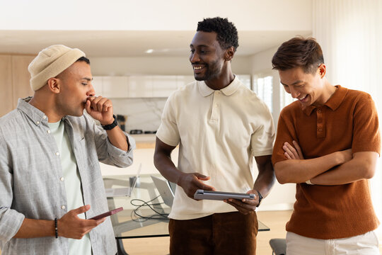 Diverse male friends laughing in kitchen in beanie and polos, holding tablet smartphone laptop